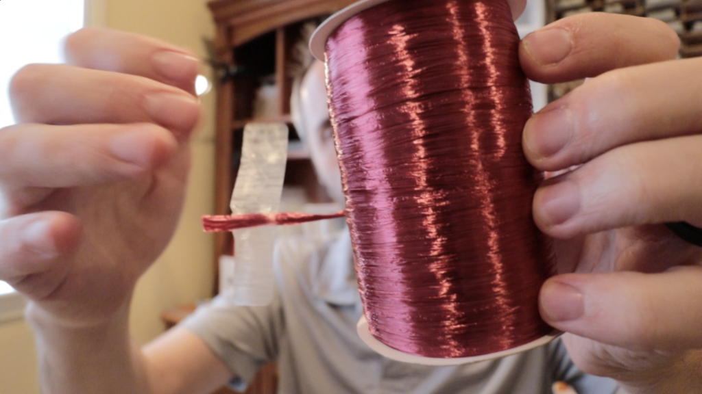 The photo features a spool of Berwick Offray Burgundy Red Pearlized Raffia Ribbon. The ribbon has a shiny, reflective surface, adding to its pearlized appearance. It's 1/4 inch wide and the spool contains 100 yards of ribbon. The burgundy red color is deep and rich, suitable for decorative purposes such as gift wrapping, crafts, or floral arrangements. The person holding the spool is displaying both the overall size and the texture of the ribbon. The surroundings suggest a home or crafting environment.